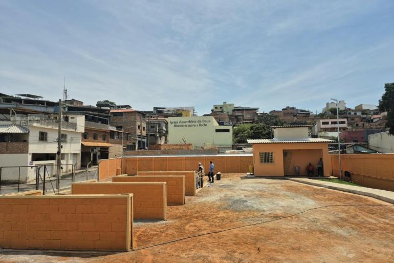 Prefeitura conclui obras da URPV São Tomás, na Região Norte A foto mostra uma área pública aberta, com piso de terra e trechos pavimentados, cercada por muros baixos pintados de laranja. Ao fundo, há uma área residencial densamente ocupada, com casas de diferentes tamanhos e estilos, muitas construídas em alvenaria aparente, distribuídas por uma encosta. No centro da imagem, algumas pessoas circulam pelo espaço, enquanto, à direita, há uma pequena edificação de cor alaranjada, com portas e janelas protegidas por grades.