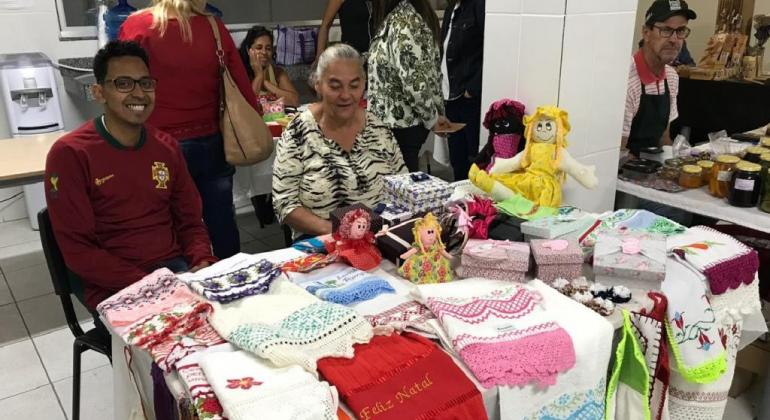 Rapaz e senhora expõem panos de pratos decorados com crochê na Mostra de Economia Popular Solidária.