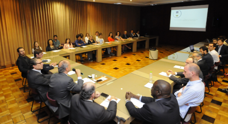 O Governo de Valonia, na Bélgica, mostrou o programa de voluntários da região belga que é parceiro Programa Municipal de Voluntariado da PBH. Foto: Rodrigo Clemente/PBH Mesa em formato de "u" com cerca de 30 pessoas em conferência, em uma sala, com telão ao fundo.