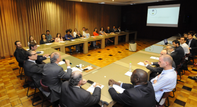 Mesa em formato de "u" com cerca de 30 pessoas em conferência, em uma sala, com telão ao fundo. 