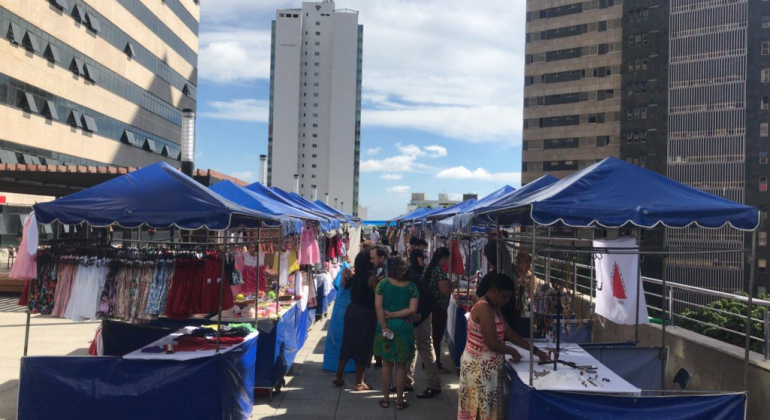 Nove barracas de lona azul e mercadorias com pessoas passando, durante o dia. 