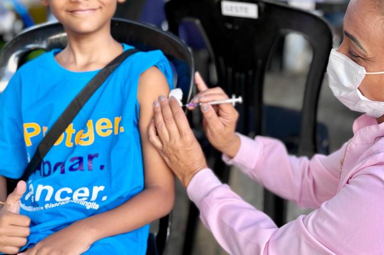 Prefeitura de BH aplicou cerca de 50 mil doses de imunizantes na Campanha de Multivacinação Criança sorridente, de camiseta azul, recebe uma injeção no braço enquanto faz sinal de positivo com a mão. Uma profissional de saúde, de máscara e blusa rosa, aplica a vacina. Cena de vacinação ao ar livre.