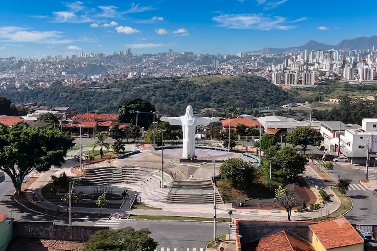 Praça no Barreiro vista de cima. Cristo com os braços abertos no centro da praça, ao seu redor algumas árvores, casas e ruas. No plano de fundo a cidade de Belo Horizonte. 