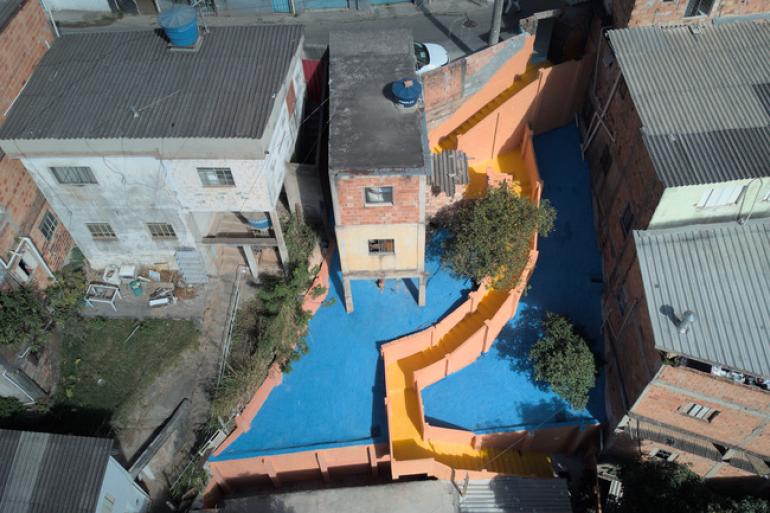 PBH conclui obra de contenção de encosta e amplia segurança na Vila Nossa Senhora de Fátima Imagem aérea de trecho reformado no Aglomerado da Serra. Escadaria pintada de amarelo comparte o pátio pintado de azul.