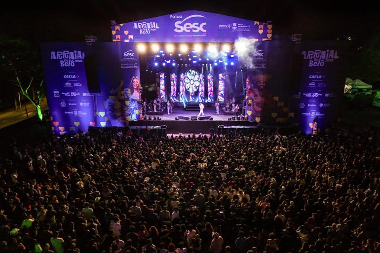 Multidão em frente ao palco. Estrutura grande e bem iluminada nas cores rosa e roxa, com palco no centro. 