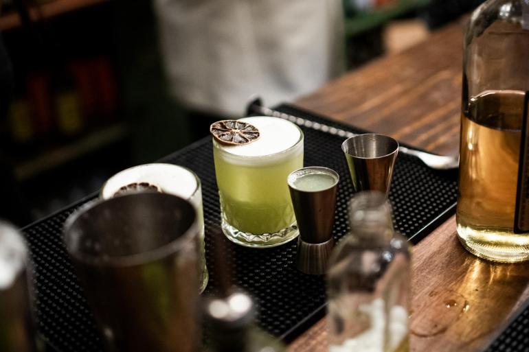 Coquetéis sendo preparados sobre o balcão de um bar de Belo Horizonte, com utensílios de preparo e garrafas de bebidas ao fundo