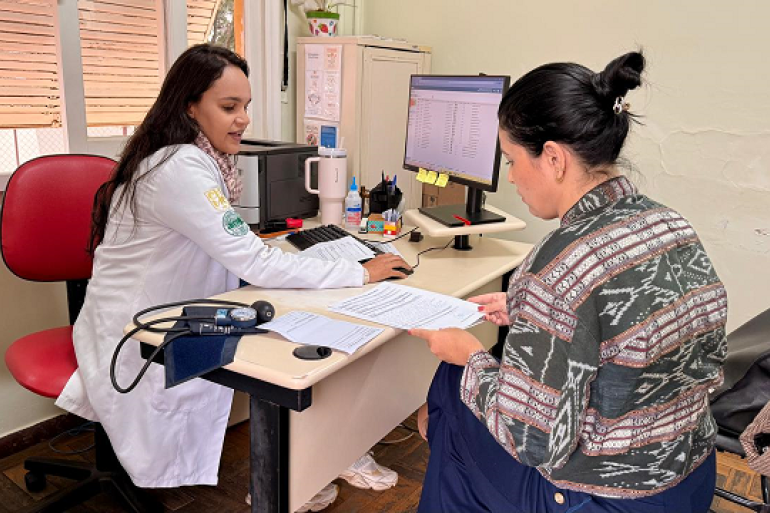 Uma profissional, vestindo jaleco branco, está sentada diante do computador e orienta uma paciente que segura alguns documentos. Sobre a mesa há materiais de trabalho e um aparelho para aferição da pressão arterial.