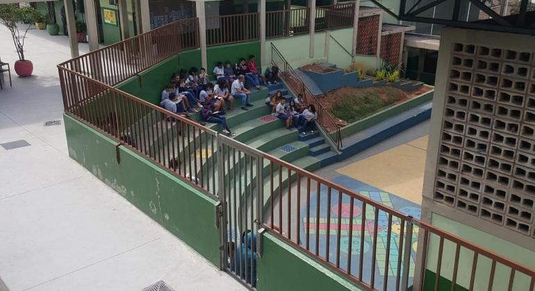 Obras possibilitam aumento de vagas nas escolas da regional Nordeste. Foto: Divulgação/PBH Pátio de escola com grades, jardim e arquibancadas com dezenas de alunos sentados.