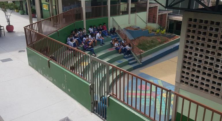 Pátio de escola com grades, jardim e arquibancadas com dezenas de alunos sentados.