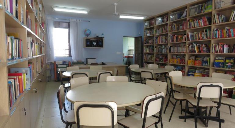 Biblioteca com mesas, cadeiras, estantes com livros, aparelho de som e equipamentos de estudo.