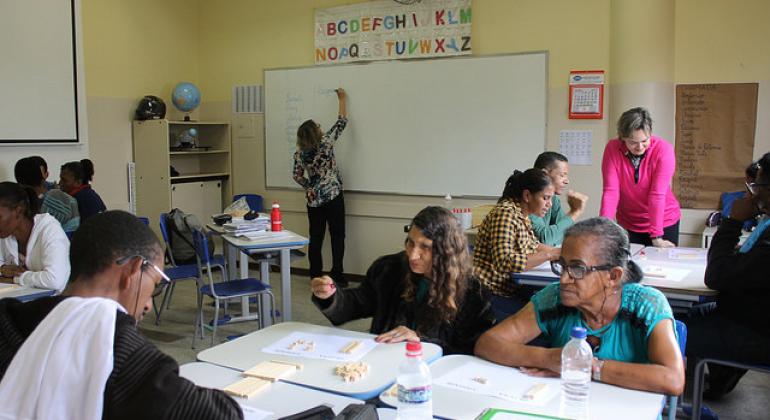 Duas professoras dão aulas para cerca de oito jovens e adultos sentados em mesas, em sala de aula.