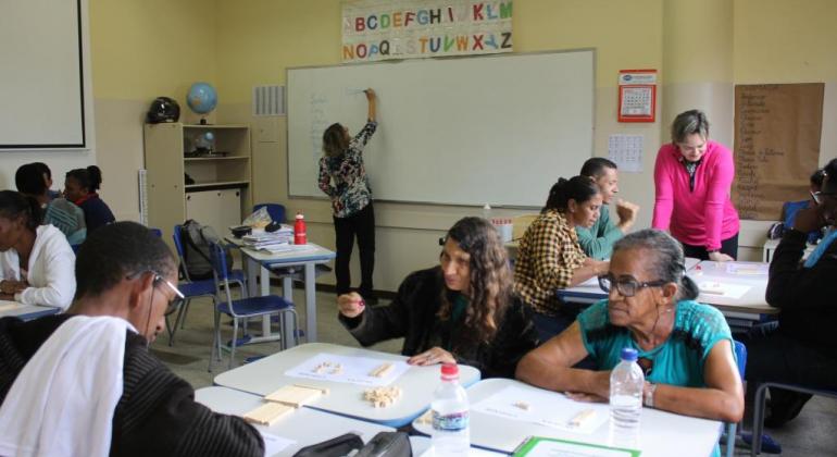 Dois professores em sala de aula de cerca com seis adultos estudando em pares.