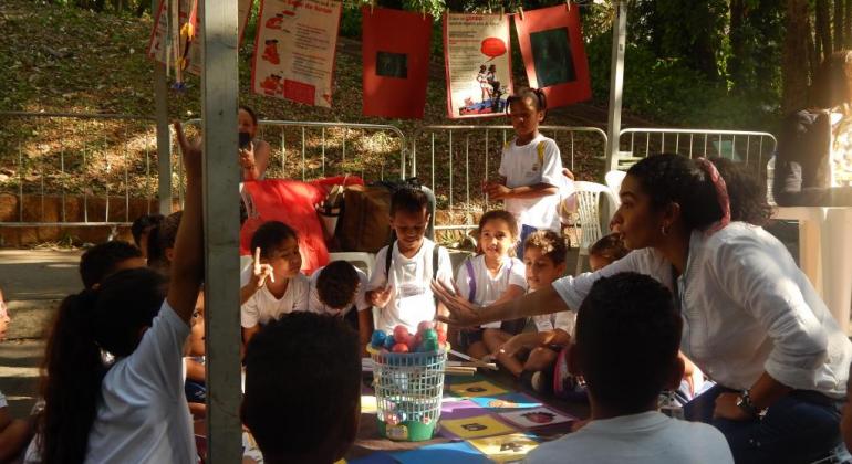 Mais de dez crianças brincam com bolinhas em um cesto em local aberto, durante o dia, acompanhadas por adulta. 