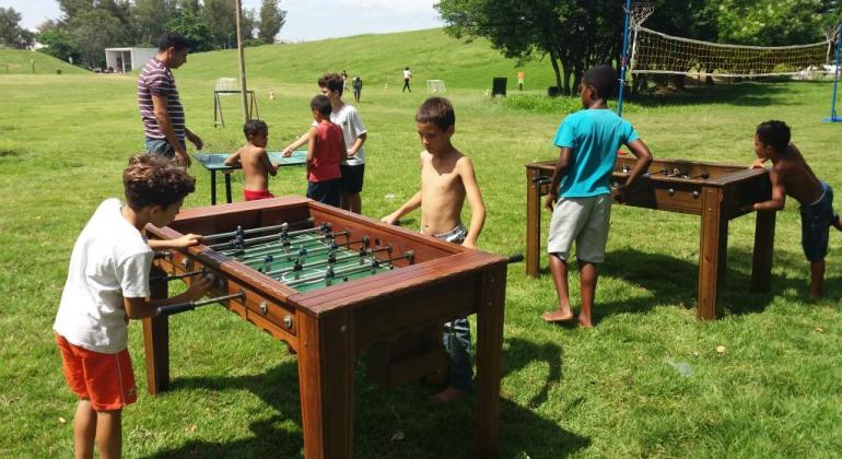 Brincar em camas elásticas, jogar totó e aprender a praticar malabarismo são algumas das novidades do programa BH em Férias, no dia 16/7. Foto: Divulgação PBH Sete crianças jogam pebolim em duas mesas em um parque gramado, a céu aberto, durante o dia.