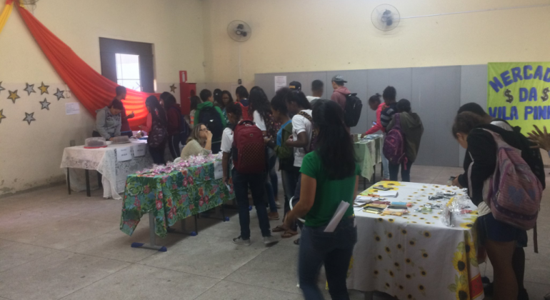 Alunos do 9º ano do Ensino Fundamental da E. M. da Vila Pinho aprenderam na prática o que é matemática e planejamento financeiro. Foto: Divulgação PBH Mais de quinze alunos observam pertences diversos em uma mesa.