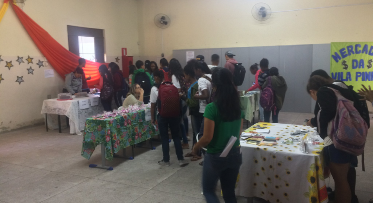 Mais de quinze alunos observam pertences diversos em uma mesa. 