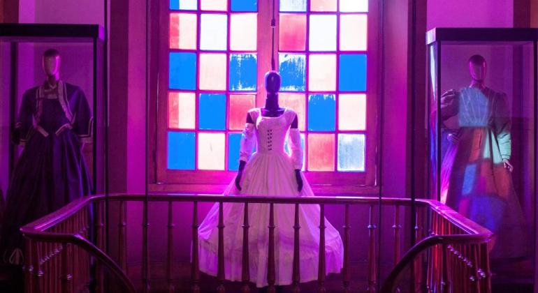 Três vestidos de época, longos e de manga comprida, um azul, um branco e um cinza, expostos entre o guarda-corpo da escadaria do Museu da Moda e os vitrais da janela, sob uma luz rósea.