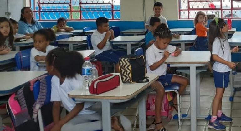 Oito alunos, sentados em suas carteiras, em sala de aula. 