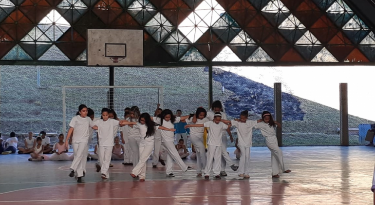 Mais de quinze crianças de branco dançam de maneira coordenada em ginásio. 
