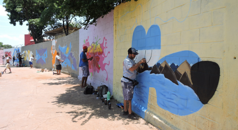 Os muros externos da Escola Municipal Professora Alice Nacif, na Pampulha, ganharam desenhos e letras coloridas em uma intervenção coletiva. Foto: Andrea Moreira/PBH Cinco jovens grafitam diferentes partes de um muro, cada uma delas com uma cor de fundo, durante o dia.