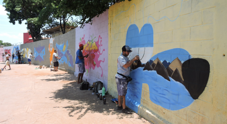 Cinco jovens grafitam diferentes partes de um muro, cada uma delas com uma cor de fundo, durante o dia. 