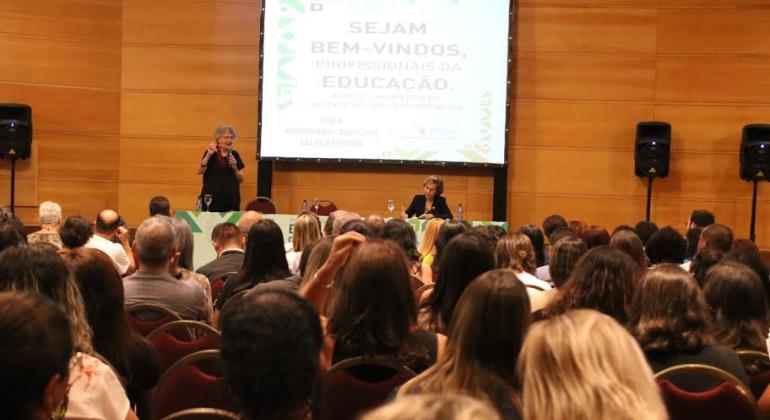 A secretária municipal de Educação, Ângela Dalben, sentada, em frente à mesa, e uma mulher palestram à frente de um telão. Mais à frente, mais de trinta pessoas assistem, sentadas. 