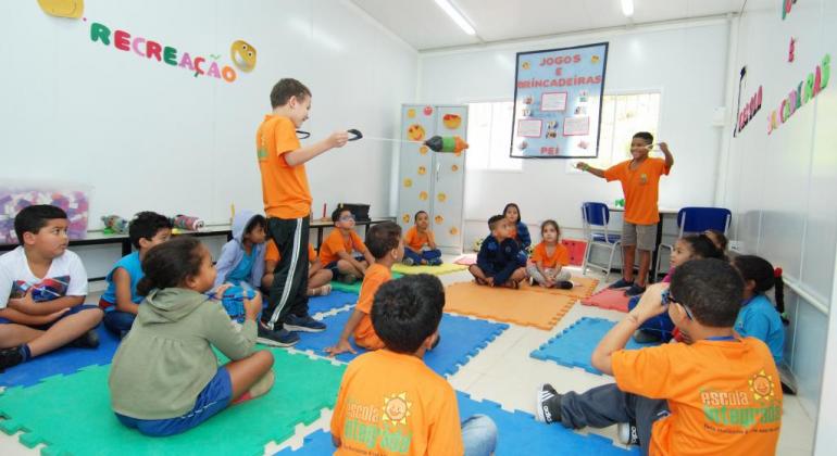 Doze alunos da escola integrada sentados em círculo, no meio deles, de pé, dois alunos jogam o jogo vai e vem, em que cada um segura duas cordas; ao abri-las com os braços, uma bola, pela qual as cordas atravessam, vai de uma lado para o outro.