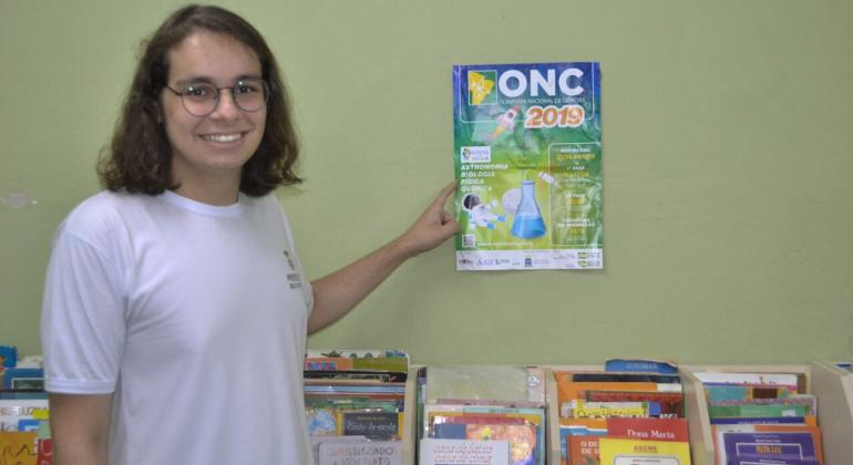 O estudante da rede de ensino municipal, Paulo César Máximo Santos Júnior, recebe na quinta-feira, dia 28/11, a medalha de ouro na Olimpíada Nacional de Ciências. Foto: Divulgação PBH Estudante aponta para cartaz na parede.