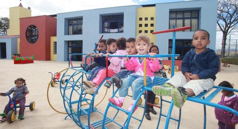 Sete crianças brincam em playground a frente de Unidade de Municipal de Educação Infantil, durante o dia. 