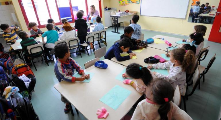 Cerca de dezessete alunos da Educação Infantil, em sala de aula, com professora ao fundo. 