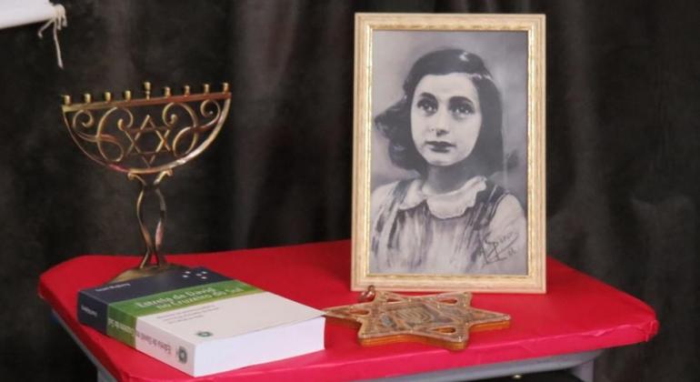Foto de Anne Frank ao lado de livro, estrela de david e chanuquiá, o candelabro judaico, sobre uma mesa de forro vermelho. Foto ilustrativa. 