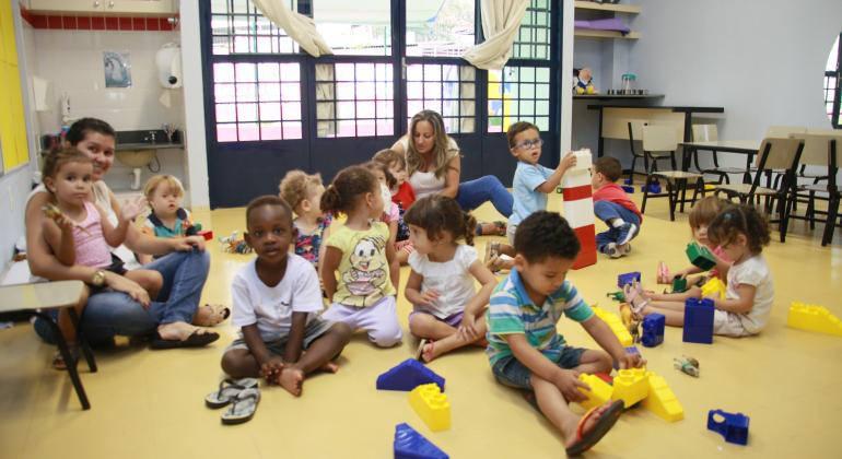 Foto: Divulgação PBH Sete crianças da Educação Infantil brincam no chão, acompanhadas por duas mulheres.