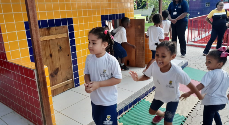 A PBH publica, no dia 30/11, a prorrogação do prazo para o cadastro de alunos com interesse no atendimento em tempo integral. Foto: Divulgação PBH Cinco crianças da escola em tempo integral brincam no pátio, acompanhada por duas mulheres, durante o dia.