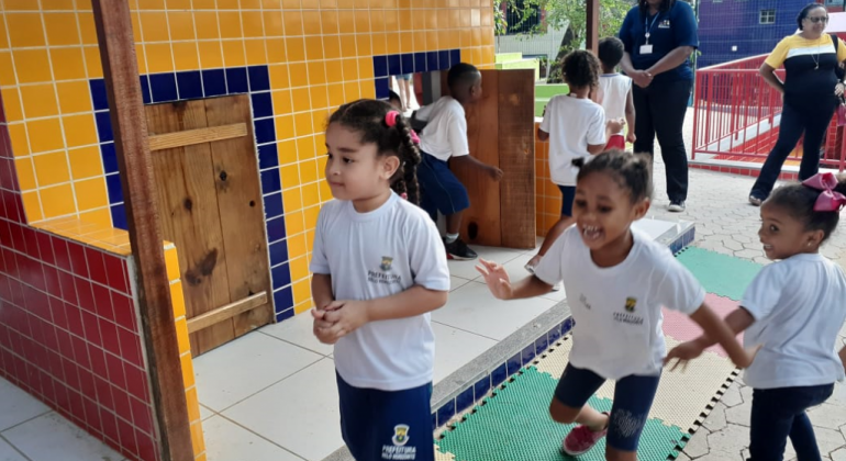 Cinco crianças da escola em tempo integral brincam no pátio, acompanhada por duas mulheres, durante o dia. 