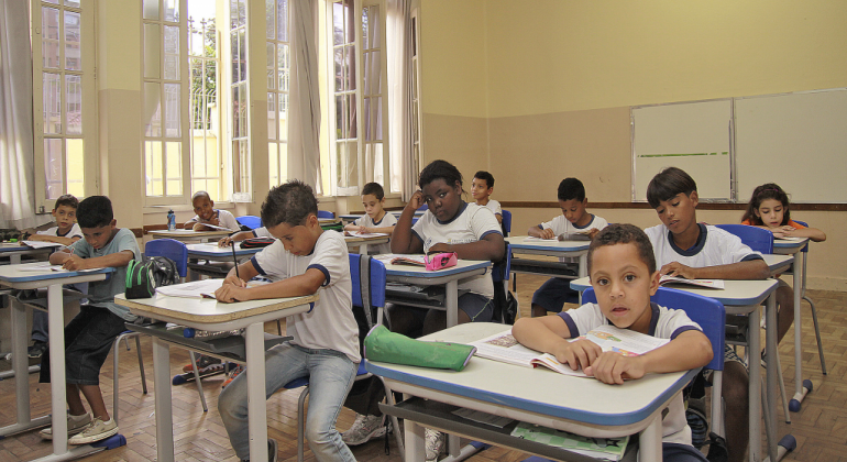 Nove crianças em sala de aula, sentadas em carteiras, durante o dia. 