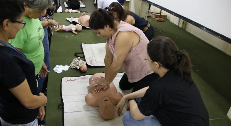 Para capacitar profissionais da educação para atuarem em situações de emergência, a PBH está promovendo cursos básicos de primeiros socorros. Foto: Divulgação PBH Mulher simula passagem cardíaca em boneco com tórax similar ao humano, acompanhada de outras três mulheres, em uma sala.