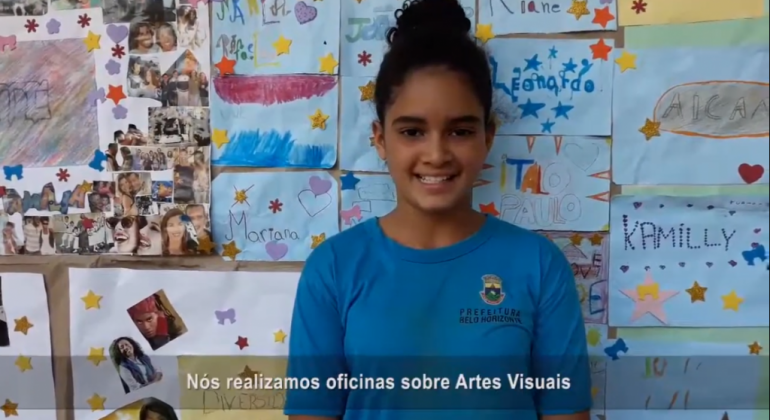 Estudante à frente de uma painel feito por alunos com legenda que diz: "Nós realizamos oficinas sobre Artes Visuais", cena de vídeo documentário Amabilidade da E. M. Santos Dumond. 