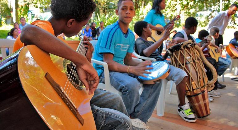 Menino toca violão, acompanhado de outras crianças que tocam pandeiro, tambor e violões, em local aberto, durante o dia. 
