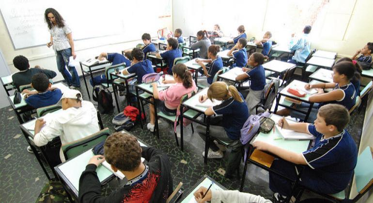 As matrículas ocorrerão entre os dias 21 e 25 de outubro nas secretarias escolares. Foto: Divulgação PBH Mais de treze alunos, sentados em suas carteiras, acompanham professora, de frente a quadro branco.