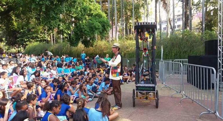 Homem de chapéu e colete colorido lê livro para mais de sessenta crianças, no parque. 