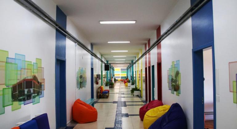 A foto mostra um corredor com paredes brancas com portas pintadas de cores variadas como azul e vermelho. Também estão colocados puffs coloridos.