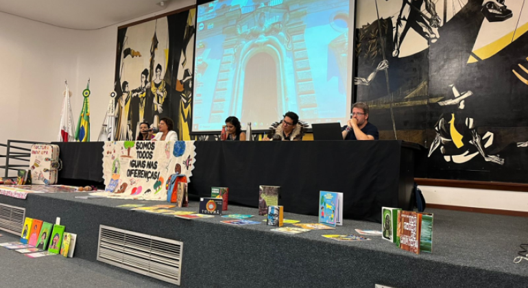 fotografia colorida de uma mesa, composta por cinco professores(as). A mesa é decorada com livros e uma faixa, na qual está escrito: “Somos todos iguais nas diferenças!”