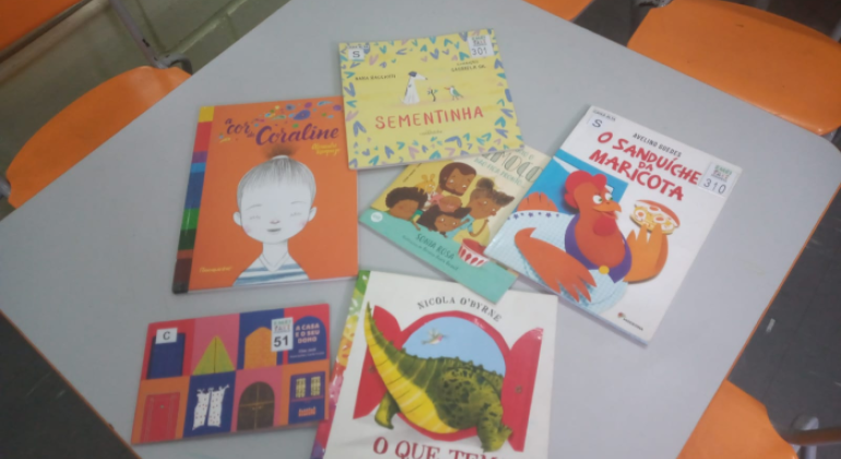 #paratodosverem: em uma mesa cinza cercada por cadeiras laranjas, estão dispostos seis livros infantis coloridos. Entre eles, “A cor de Coraline”, “Sementinha”, “O sanduíche da Maricota”, “O que tem atrás da porta?”, “O que tem atrás do medo?” e “A casa e o seu dono”. As capas trazem ilustrações vibrantes de animais, pessoas e cenários lúdicos.