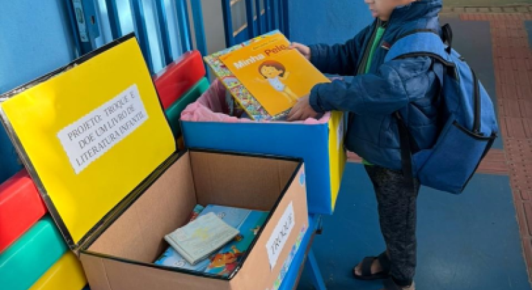 #paratodosverem: Um menino de mochila azul e casaco observa um livro infantil intitulado “Minha Pele” diante de caixas de papelão sobre um banco colorido. As caixas têm placas com os dizeres “PROJETO: TROQUE E DOE UM LIVRO DE LITERATURA INFANTIL” e “TROQUE”. Dentro delas há outros livros disponíveis para troca.