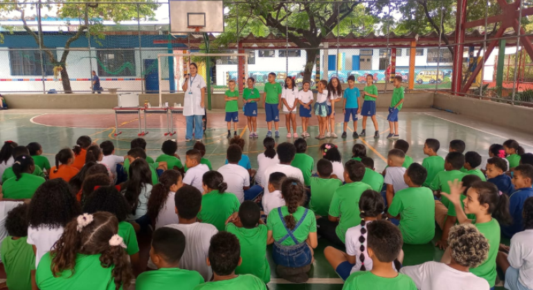 #paratodosverem: em uma quadra escolar coberta, diversas crianças sentadas, vestidas com uniforme verde, assistem a uma apresentação. À frente, uma profissional de jaleco branco está ao lado de uma mesa com materiais e de um grupo de estudantes. 
