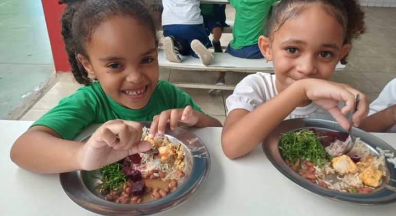 #paratodosverem: a imagem mostra duas meninas sorrindo enquanto fazem uma refeição em um ambiente escolar. Elas estão sentadas à mesa com pratos de comida equilibrada, contendo arroz, feijão, vegetais como couve e beterraba, além de pedaços de frango. A menina à esquerda veste uma camiseta verde, enquanto a menina à direita usa uma blusa branca e tem um laço azul no cabelo. Ao fundo, outras crianças também estão se alimentando.