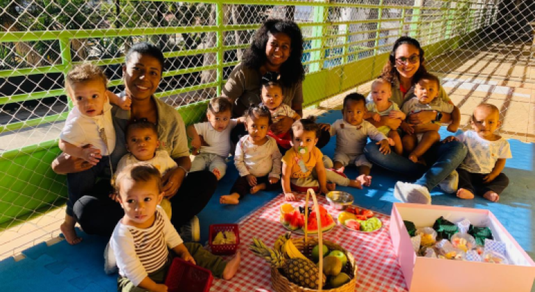 #paratodosverem: A imagem mostra três mulheres sentadas no chão com um grupo de bebês sobre um tapete azul. À frente, há uma toalha xadrez vermelha e branca com frutas como melancia, banana, abacaxi e kiwi, além de uma caixa rosa com lanches. O local é cercado por uma grade verde e está iluminado pela luz do sol.