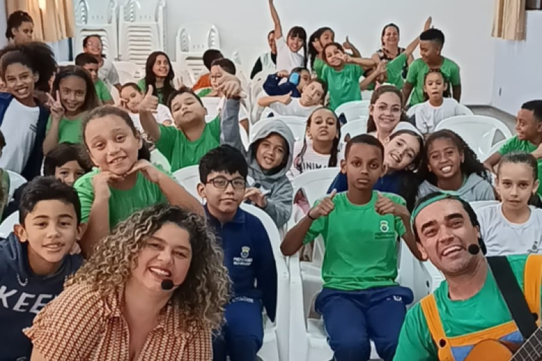 #paratodosverem: Em um auditório com cadeiras brancas, um grande grupo de crianças e adultos está posando para a foto. A maioria sorri, faz "joinha" ou levanta a mão, demonstrando entusiasmo. Em primeiro plano, uma mulher de cabelo cacheado e um homem tocando violão olham para a câmera. A cena é alegre e passa a sensação de um evento ou apresentação de sucesso.
