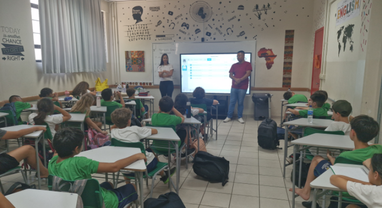 #paratodosverem: a imagem mostra uma sala de aula, com decoração temática da cultura pop. Os(as) alunos(as) estão uniformizados(as) de verde e branco e sentados em carteiras escolares organizadas em fileiras. As crianças estão atentas à apresentação feita por profissionais à frente da sala. Uma mulher, vestindo blusa listrada e calça preta, está com os braços cruzados ao lado de um homem de camiseta vermelha e jeans, que parece estar falando. Atrás de ambos, projeta-se uma apresentação.