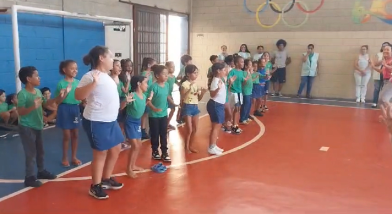 #paratodosverem: em uma quadra, duas fileiras de estudantes se apresentam para o restante da escola. Atrás delas, há uma parede pintada de azul, em que outras crianças estão encostadas, assistindo, sentadas, à apresentação dos(as) colegas. À direita, em frente a uma parede com desenhos coloridos, incluindo os anéis olímpicos, pessoas adultas observam a atividade. 